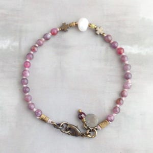 May include: A delicate bracelet featuring small, round, pink and purple beads strung together. Accents include gold-toned metal flower-shaped beads, a white pearl, and a decorative clasp. The bracelet is designed for a comfortable fit.
