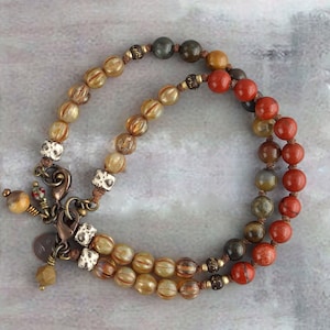 Op de afbeelding: Een meerstrengige armband met bruine, rode en grijze kralen. De armband heeft een sluiting en een decoratieve hanger.