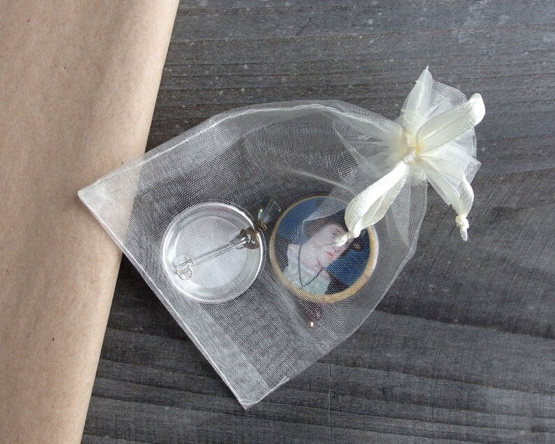 May include: A white organza bag with a white satin ribbon bow, containing a round button with a silver pin and a round button with a portrait of a woman in a blue dress.