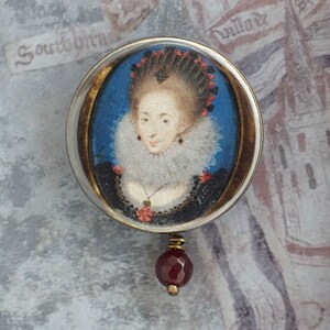 May include: A round, silver-toned locket with a portrait of a woman in a white ruff and black dress. The locket has a red gemstone pendant.