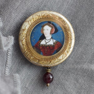May include: A gold-toned round brooch with a portrait of a woman in a red dress and a blue and white headdress. The brooch has a burgundy red gemstone pendant.