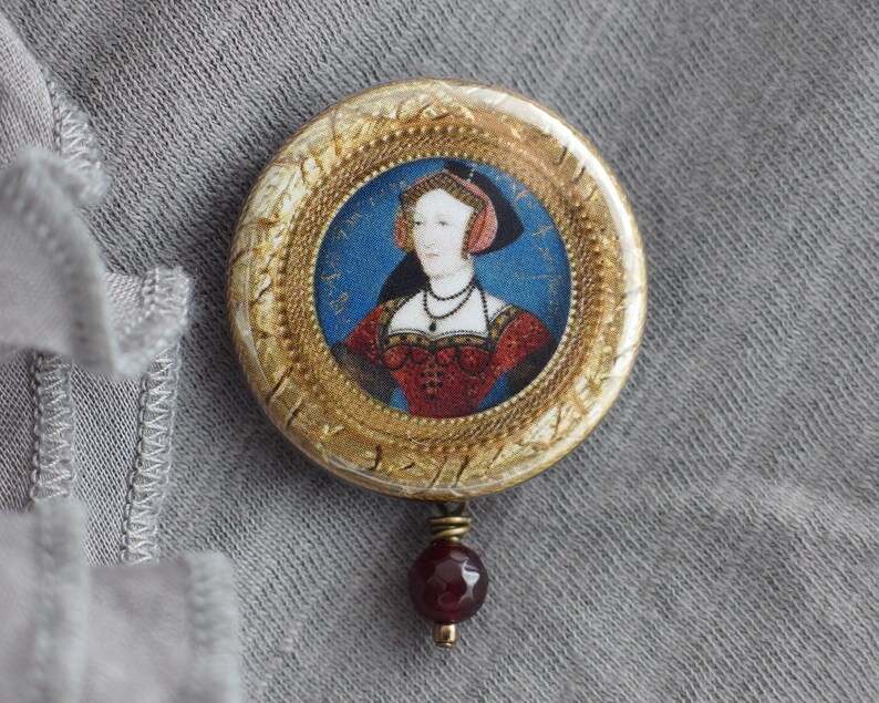 May include: A gold-toned round brooch with a portrait of a woman in a red dress and a blue and white headdress. The brooch has a burgundy red gemstone pendant.