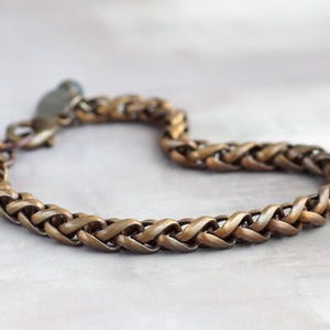 Op de afbeelding: Een bronskleurige armband met een gevlochten kettingontwerp. De armband heeft een karabijnsluiting. Het metaal heeft een licht verouderd uiterlijk, met kleurvariaties. Het is een sieraad.