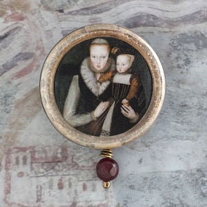 May include: A silver-toned pendant with a round glass cameo featuring a painted portrait of a woman and a child. The cameo is surrounded by a silver-toned frame. A red gemstone hangs from the bottom of the pendant.