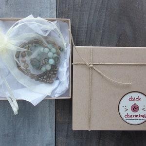 May include: A brown cardboard box with a string tied around it. The box has a sticker on it that says "chick & charming" with a small ampersand symbol. The box is sitting on a wooden surface. Inside the box is a small white bag with a green and brown beaded necklace.