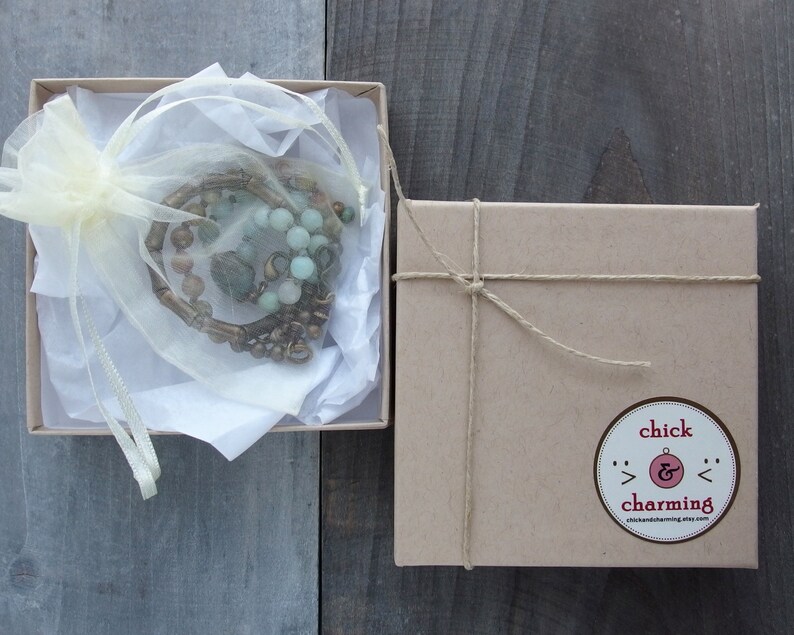 May include: A brown cardboard box with a string tied around it. The box has a sticker on it that says "chick & charming" with a small ampersand symbol. The box is sitting on a wooden surface. Inside the box is a small white bag with a green and brown beaded necklace.
