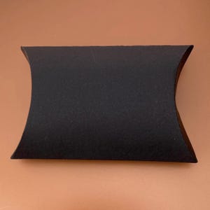 Puede incluir: Una caja negra en forma de almohada con bordes curvos. La caja está hecha de un material texturizado y tiene forma rectangular. La caja está vacía y lista para ser llenada con pequeños regalos o productos.
