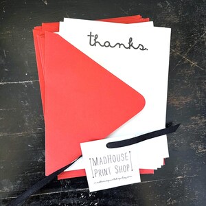 May include: A stack of white cards with the word "thanks" in black script, paired with red envelopes. A small white tag with "MADHOUSE PRINT SHOP" is attached with a black ribbon. The cards and envelopes are on a dark wooden surface.