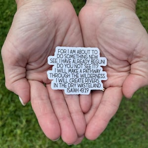 May include: A white rectangular sticker with black text, held in open hands against a green background. The text reads "FOR I AM ABOUT TO DO SOMETHING NEW...ISAIAH 43:19". The sticker is a religious or inspirational item.