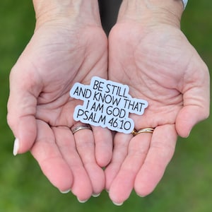 May include: A white sticker with the text "BE STILL AND KNOW THAT I AM GOD. PSALM 46:10" is held in open hands. The sticker has a light blue border and black text. The background is blurred.