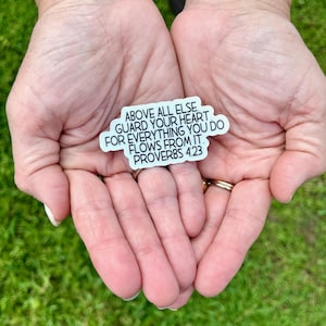 May include: A white sticker with black text, held in open hands against a green background. The sticker reads "ABOVE ALL ELSE GUARD YOUR HEART. FOR EVERYTHING YOU DO FLOWS FROM IT. PROVERBS 4:23."