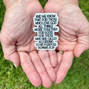 May include: A white sticker with black text, held in open hands. The sticker features the quote "AND WE KNOW THAT FOR THOSE WHO LOVE GOD ALL THINGS WORK TOGETHER FOR GOOD FOR THOSE WHO ARE CALLED ACCORDING TO HIS PURPOSE ROMANS 8:28."