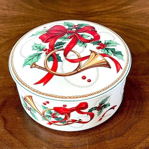 May include: A white ceramic trinket box with a Christmas theme. The lid features a red bow, holly leaves, and gold horns. The box is decorated with a white pearl rope and a green rope.