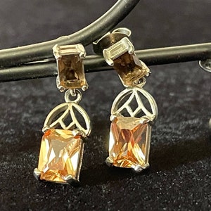 May include: A pair of dangle earrings with a gold-toned metal setting and a rectangular amber-colored gemstone. The earrings are hanging from a black jewelry stand.