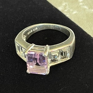May include: A silver ring with a light purple square-shaped gemstone surrounded by smaller clear gemstones. The ring is marked "925" on the inside band.