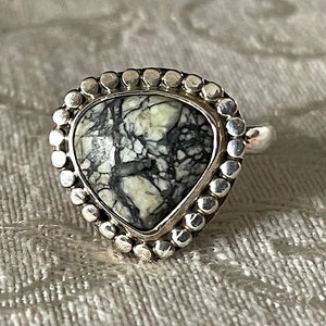 Può includere: Un anello d'argento con una pietra bianca e nera incastonata in una montatura a forma di cuore. L'anello è circondato da un bordo perlato.