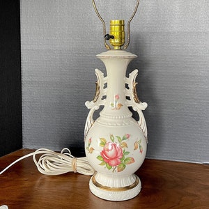 Może przedstawiać: Biała ceramiczna lampa stołowa z pozłacanymi zdobieniami i kwiatowym wzorem. Lampa ma różową różę i zielone liście na podstawie. Lampa ma złoty metalowy klosz.