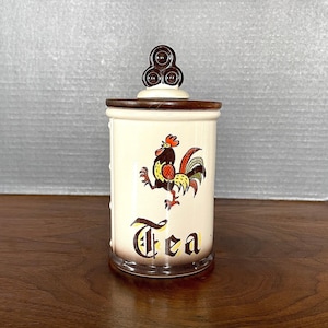 May include: A ceramic tea canister with a rooster design and the word "Tea" in a decorative font. The canister has a brown and white color scheme with a brown lid.