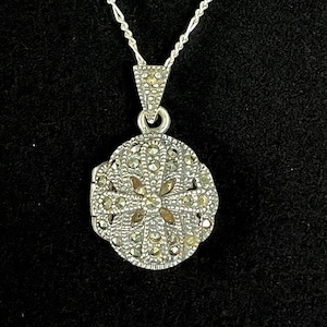 May include: A silver locket necklace with a delicate chain. The locket is oval shaped and features a floral design with marcasite stones.