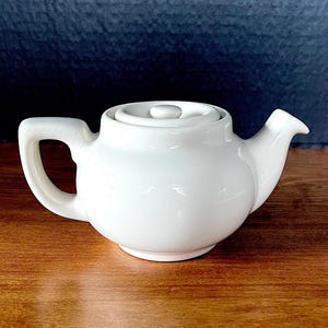 May include: A solid white ceramic teapot featuring a rounded body, a short curved spout, and a sturdy handle. The teapot includes a matching circular lid with a small central knob, presenting a smooth, glossy finish throughout the entire piece.