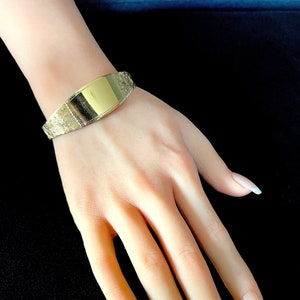 Puede incluir: Un brazalete dorado con una placa rectangular grabada en blanco. El brazalete está en la muñeca de una persona.