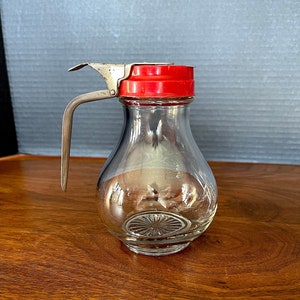Pode incluir: Um dispensador de xarope de vidro transparente com uma tampa de plástico vermelha e uma alça de metal prateada. O dispensador tem um fundo texturizado.