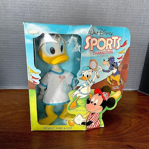 May include: A vintage Walt Disney Sports Characters doll of Daisy Duck wearing a white tennis dress with a blue trim and a white headband. The doll has yellow legs and arms and is holding a tennis racket. The doll is packaged in a box with a blue and yellow background and the text "Walt Disney Sports Characters by Durham" on the front.