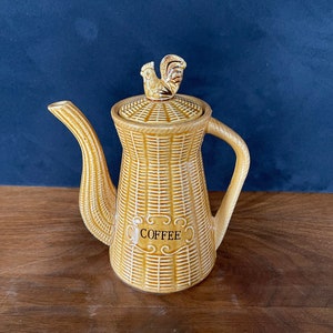May include: A brown ceramic coffee pot with a woven pattern and a rooster finial on the lid. The pot has the word "COFFEE" written on the front.