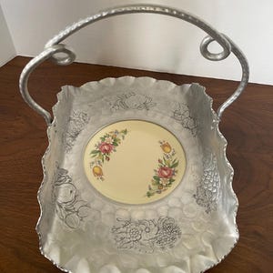 May include: A silver aluminum serving tray with a scalloped edge and a handle. The tray has a hammered texture and is decorated with embossed floral and fruit designs. A small, round, cream-colored plate with a floral design is centered in the tray.