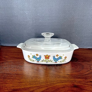 Könnte beinhalten: Weiße Auflaufform mit Glasdeckel. Die Form hat ein Blumenmuster mit blauen Vögeln und roten Blumen.