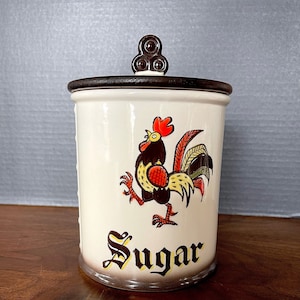 May include: A white ceramic sugar jar with a brown lid and a rooster design. The rooster is yellow, red, and brown with a black beak and comb. The jar has the word "Sugar" written in brown and yellow lettering.