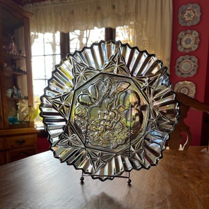 May include: A clear glass plate with a scalloped edge and a raised design of grapes and fruit. The plate is on a wooden stand.