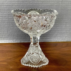 May include: A clear glass pedestal bowl with a cut glass design. The bowl has a scalloped edge and a tall, fluted pedestal.