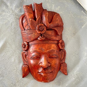May include: A wooden mask with a carved face and intricate details. The mask is painted in shades of brown and red, and features a traditional headdress.