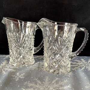 May include: Two clear glass pitchers with a diamond pattern. The pitchers have a handle on one side and a spout on the other.