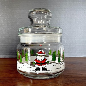 May include: A clear glass jar with a white lid and a Christmas scene painted on the side. The scene features Father Christmas, snow-covered trees, and a snowy background.
