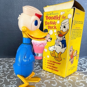 Puede incluir: Un juguete vintage de Donald Duck soplando burbujas. El pato de plástico amarillo lleva un traje de marinero azul y un gorro de marinero blanco. El pato sostiene una varita de burbujas rosa y blanca. La caja es amarilla con una imagen de Donald Duck soplando burbujas. La caja dice "WALT DISNEY'S Donald the Bubble Duck".