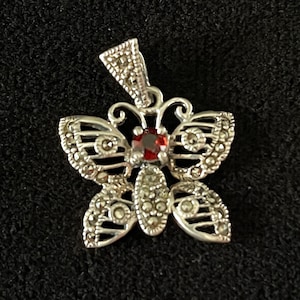 May include: Silver butterfly pendant with marcasite accents and a central red gemstone. The pendant features detailed wing patterns and a loop for a chain. The butterfly design is set against a dark background.