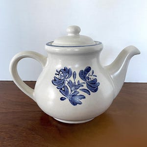 May include: A cream-colored teapot with a blue floral design. The teapot has a rounded body and a spout on the right side. The handle is on the left side.