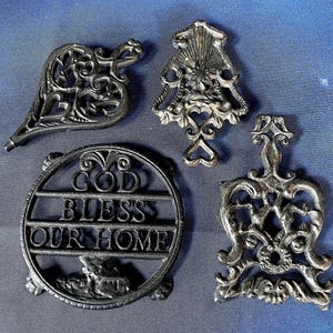 May include: Four decorative cast iron trivets in various shapes. One is heart-shaped, another is a circle with the words "GOD BLESS OUR HOME", and two others have ornate designs. The trivets are dark gray and sit on a blue surface.