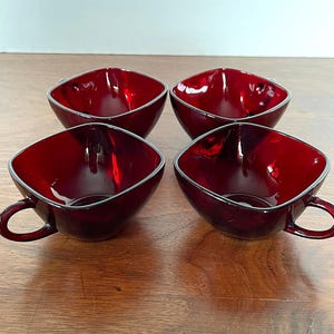 May include: Four square-shaped, red glass teacups with handles. The cups are arranged in a row on a wooden surface.