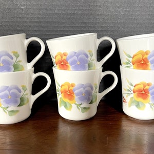 May include: Six white ceramic mugs with a floral design featuring orange, yellow, and purple pansies. The mugs are stacked in pairs.