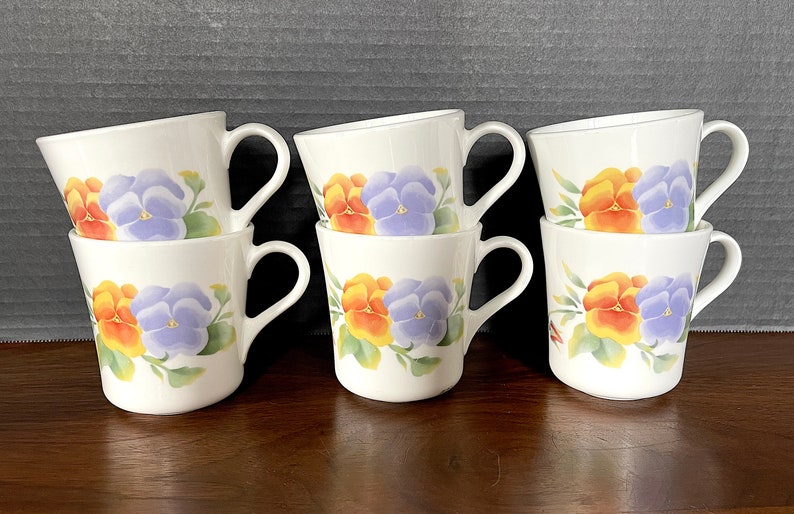 May include: Six white ceramic mugs with a floral design featuring orange, yellow, and purple pansies. The mugs are stacked in pairs.