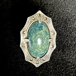 May include: A vintage silver-toned brooch with an oval, speckled blue-green stone center. The brooch has an ornate, geometric frame with small, sparkling accents. The background is black.