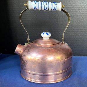 May include: A copper tea kettle with a white and blue handle and lid knob. The kettle has a spout and a curved handle. The handle and lid knob are white with blue floral designs. The kettle is made of copper and has a shiny finish.