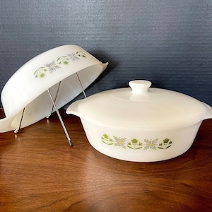 May include: A white casserole dish with a lid, both featuring a green floral pattern. The dish is on a wooden surface.
