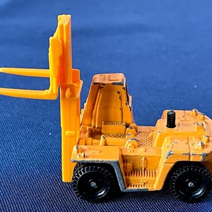 May include: A small, weathered, orange toy forklift with black wheels. The forklift has a detailed design, including a driver's compartment and lifting forks. The toy is set against a blue background.