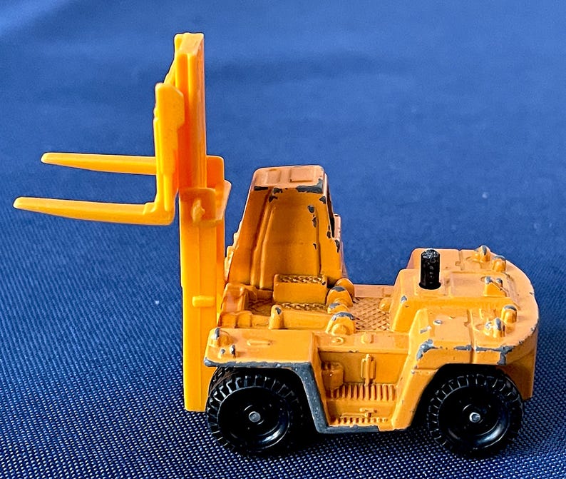 May include: A small, weathered, orange toy forklift with black wheels. The forklift has a detailed design, including a driver's compartment and lifting forks. The toy is set against a blue background.