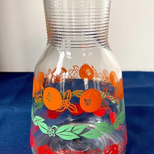 May include: Clear glass pitcher with a red lid, decorated with orange and red fruit designs. The pitcher has a ribbed neck and a wide base, perfect for serving beverages. The fruit designs include oranges, tomatoes, and green leaves.