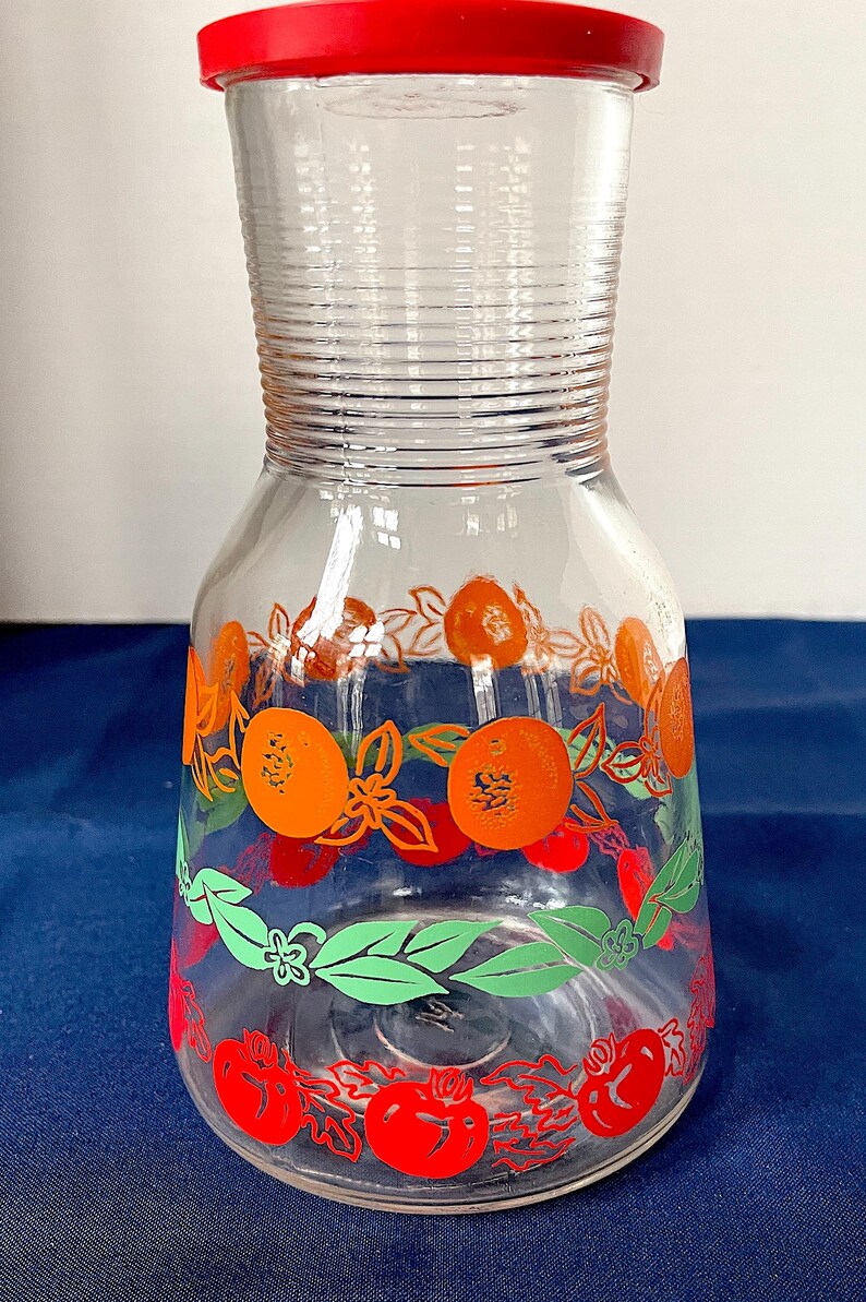 May include: Clear glass pitcher with a red lid, decorated with orange and red fruit designs. The pitcher has a ribbed neck and a wide base, perfect for serving beverages. The fruit designs include oranges, tomatoes, and green leaves.